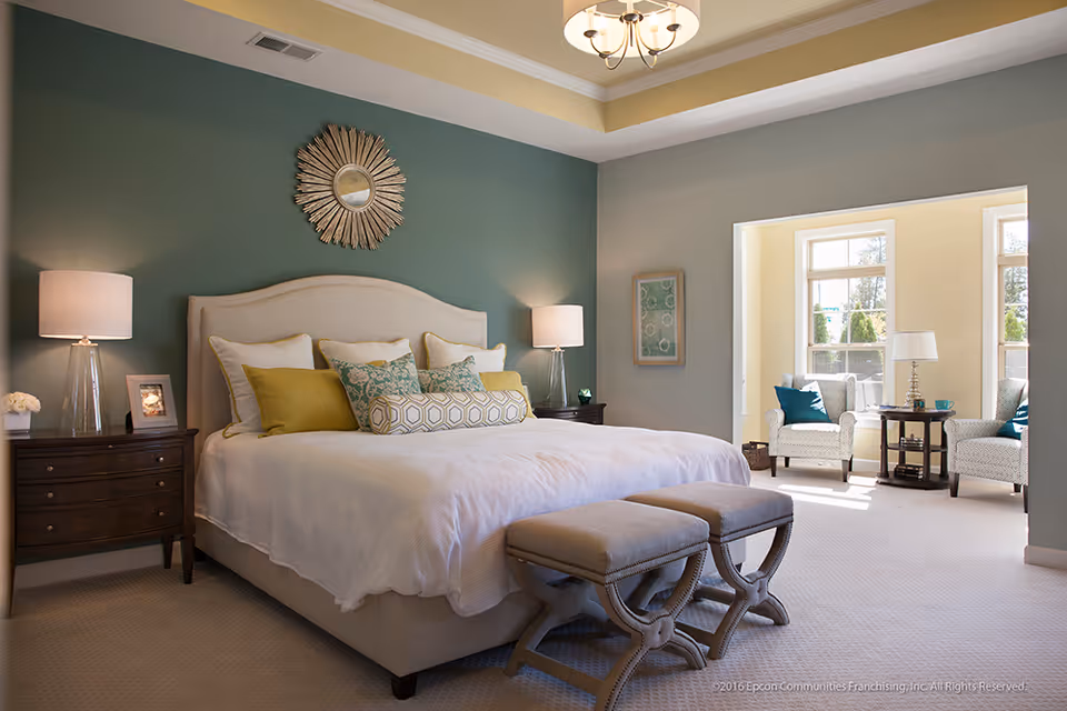 A well-lit bedroom with a large bed featuring a beige upholstered headboard, white bedding, and multiple decorative pillows in green and yellow tones. Two matching dark wood nightstands with lamps flank the bed. Above the bed is a decorative sunburst mirror. In the background, a sitting area with two patterned armchairs, a small round table, and a lamp is visible near large windows letting in natural light.