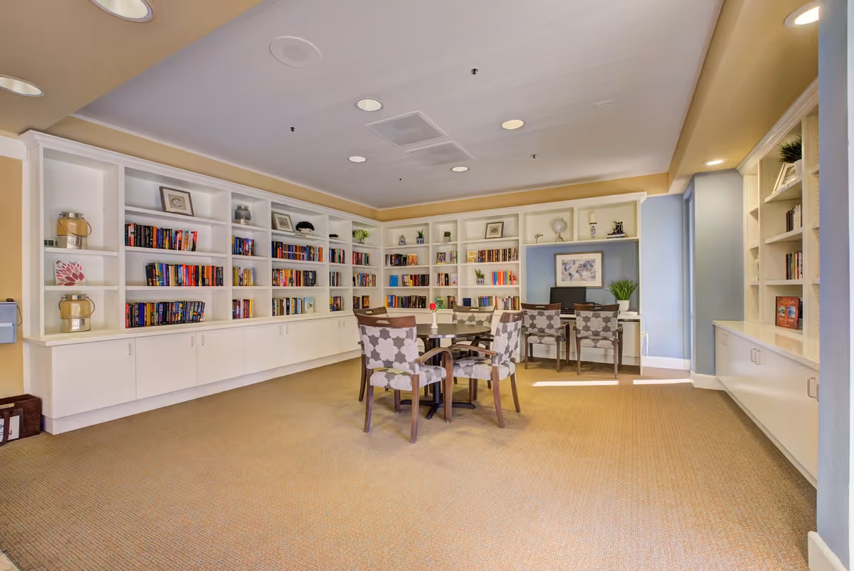 A bright and spacious room with beige carpet and light-colored walls featuring built-in white bookshelves filled with books and decorative items. In the center, there is a round table surrounded by four patterned chairs. At the back, there is a desk with two chairs, a computer, and framed artwork on the wall. The room is well-lit with recessed ceiling lights.