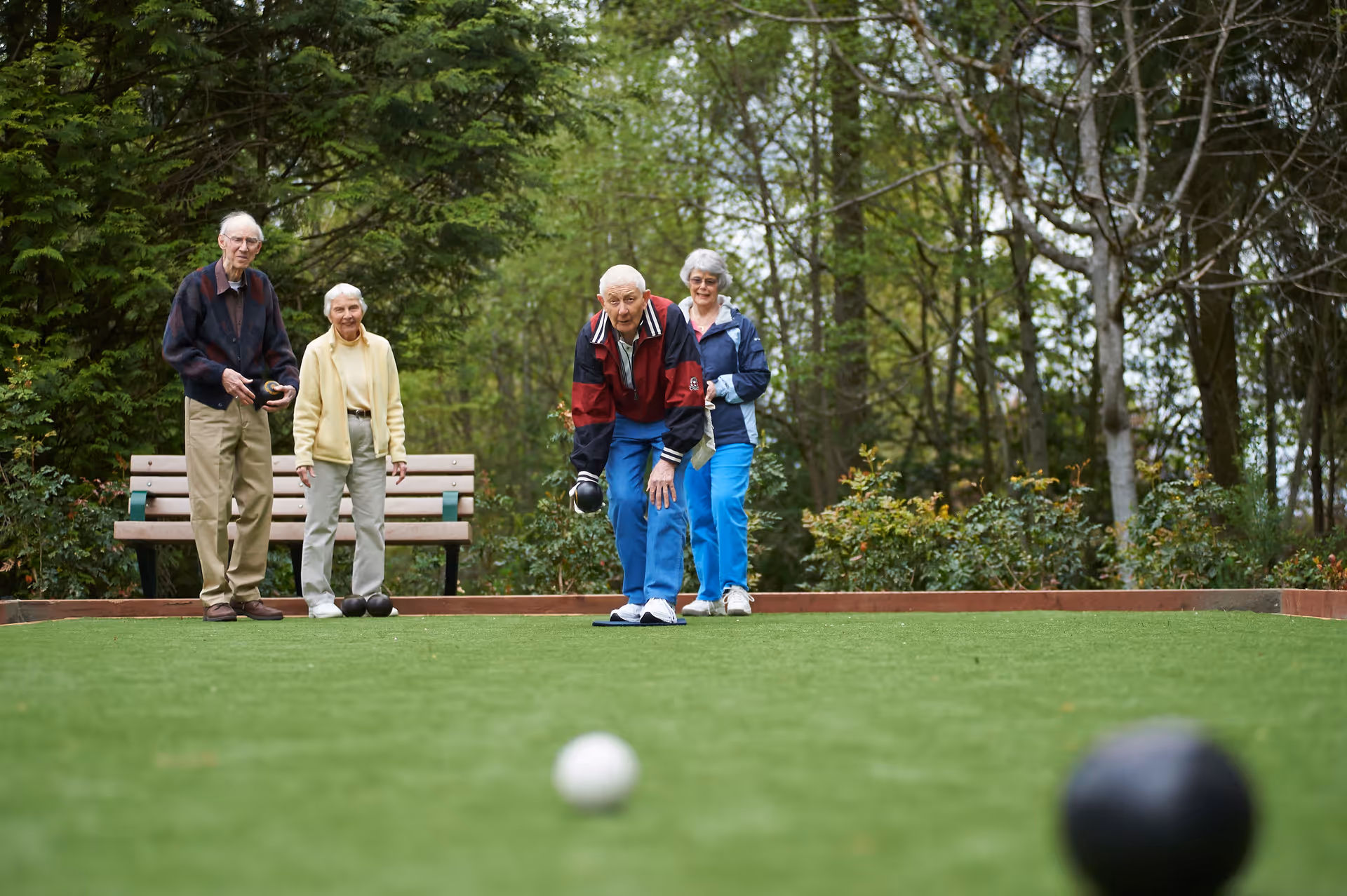 Four elderly people playing bocce ball on a green outdoor court surrounded by trees and bushes, with a wooden bench in the background.
