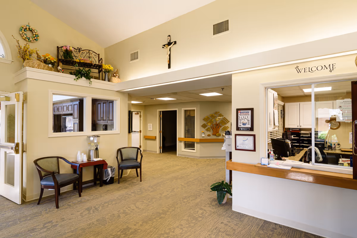 Interior view of a nursing center reception area with a welcome sign above the reception window. There are two chairs and a small table with a water dispenser and cups on the left side. The walls are decorated with plants, flowers, and a crucifix. The area is well-lit with beige walls and carpeted floors.