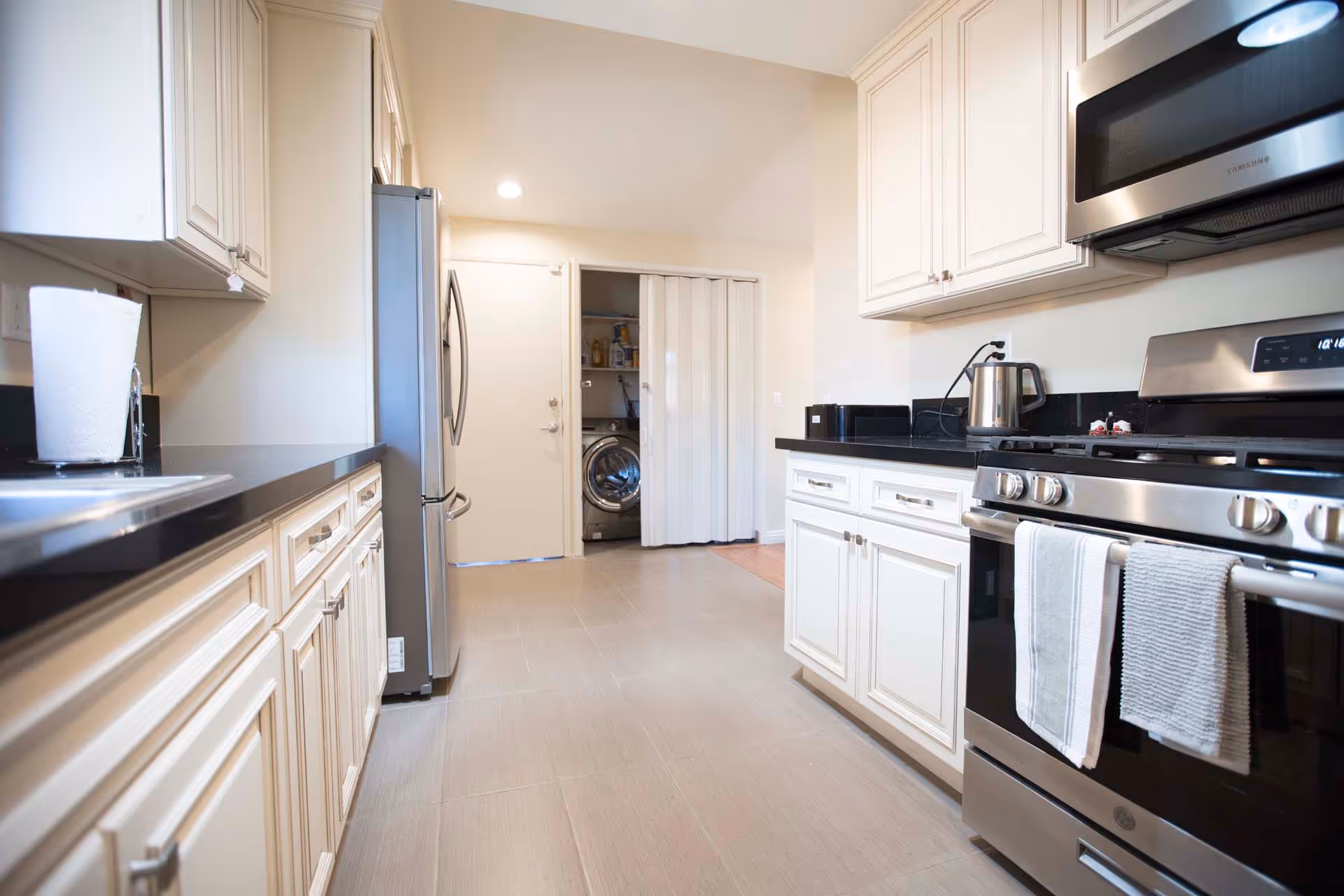 A modern kitchen with white cabinetry and black countertops. Stainless steel appliances include a refrigerator, microwave, and gas stove with two towels hanging on the oven handle. A paper towel holder is on the left countertop, and a stainless steel electric kettle is on the right countertop. At the back, there is a laundry area with a washing machine visible behind a partially open folding door.