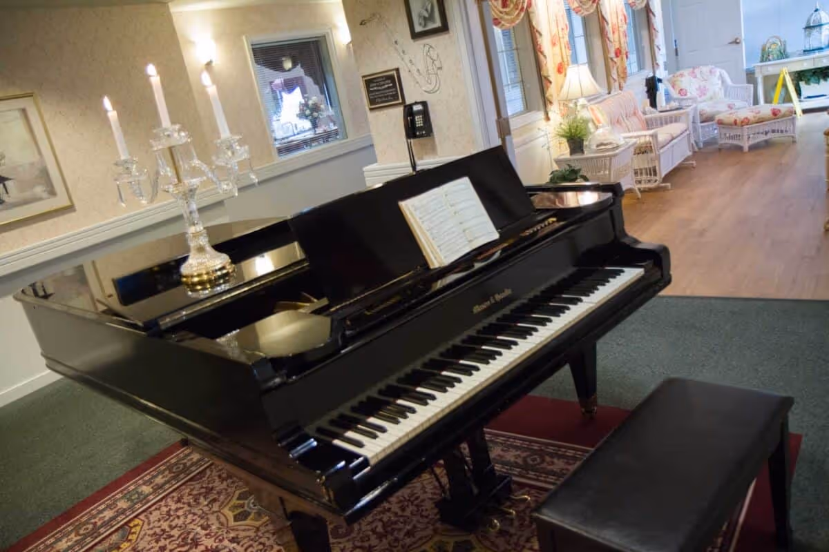 A black grand piano with an open music book on the stand, placed on a patterned rug in a cozy room. To the right, there is a seating area with white wicker furniture, floral cushions, and a wooden floor. The room has soft lighting and decorative wall art.