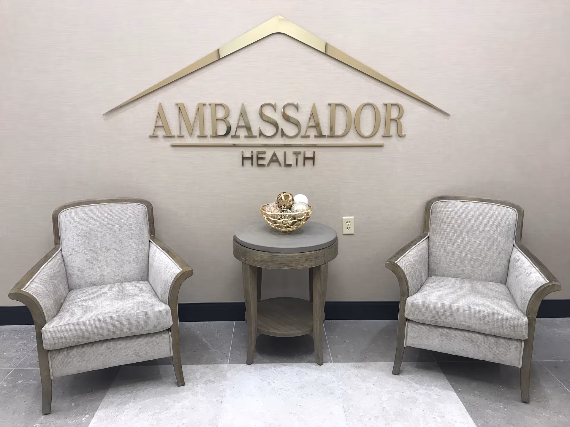 Two beige upholstered armchairs with wooden frames are positioned on either side of a small round wooden table with a decorative bowl containing ornamental spheres. Behind the furniture, a wall displays the text 'AMBASSADOR HEALTH' with a stylized roof design above the text.