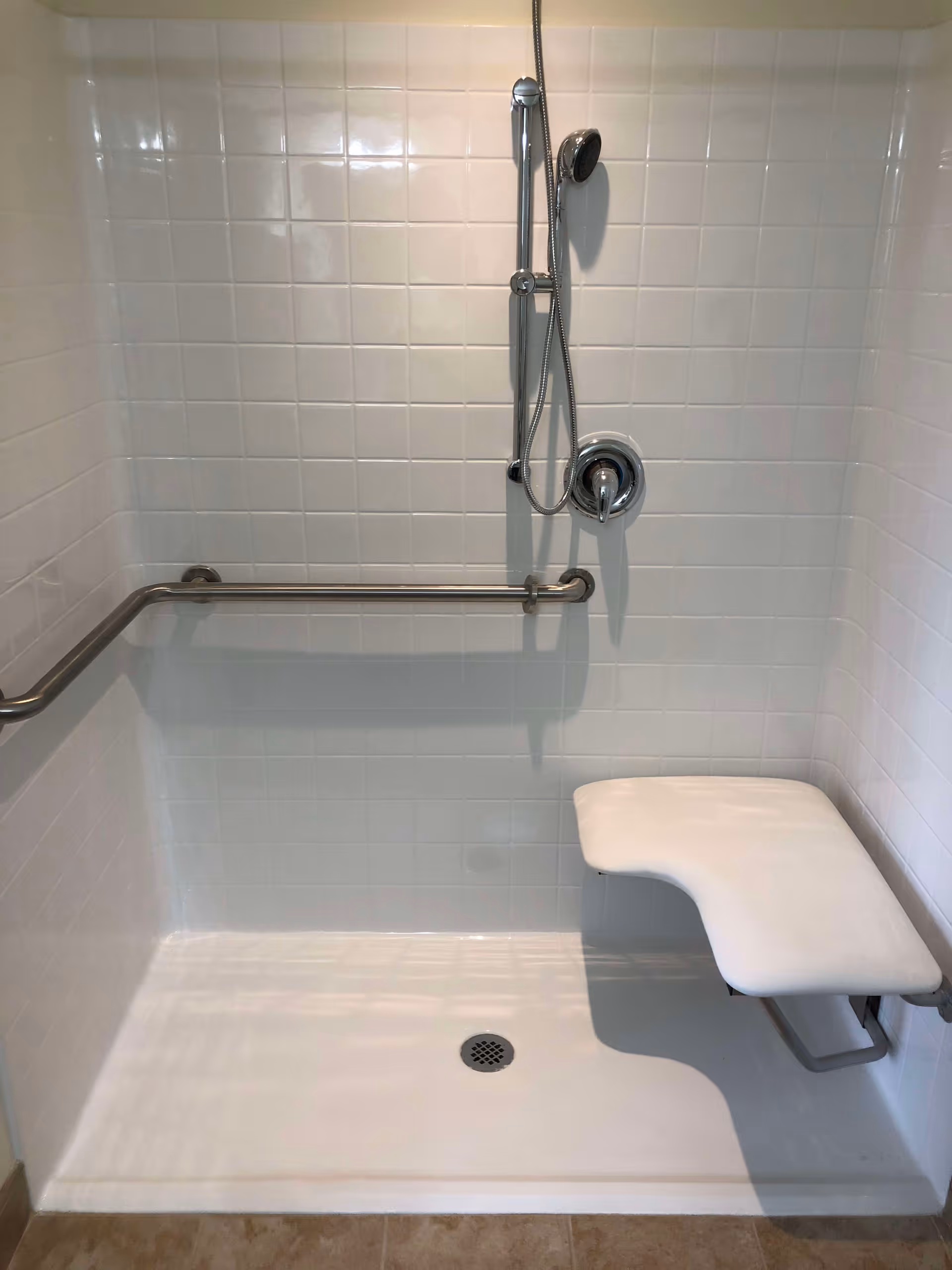 A white tiled shower area with a handheld showerhead attached to a vertical bar, a horizontal grab bar on the left wall, and a foldable white shower seat mounted on the right wall. The shower floor is white with a central drain.
