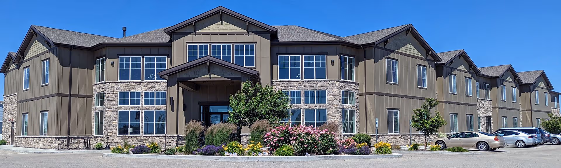 Exterior view of a two-story senior living facility building with a combination of stone and siding facade, large windows, a covered entrance, landscaped garden with colorful flowers and shrubs, and a parking lot with several cars under a clear blue sky.