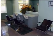 Interior view of a salon area with two black salon chairs in front of a counter with a large mirror. There is a flower arrangement on the counter and a black chair to the right side. The room has light-colored walls and windows with blinds.