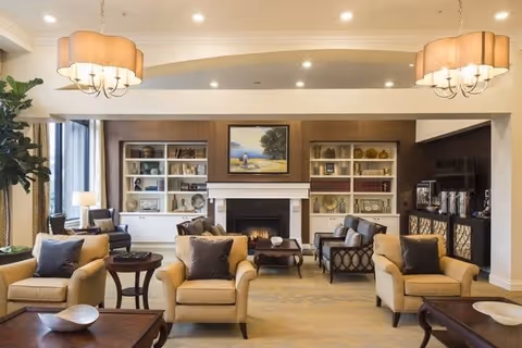 A spacious and well-lit living room with beige armchairs and sofas arranged around a central coffee table. The room features a fireplace with a painting above it, built-in white bookshelves filled with decorative items and books, and two large pendant lights hanging from the ceiling. There are side tables with lamps and a large flat-screen TV on the right side.