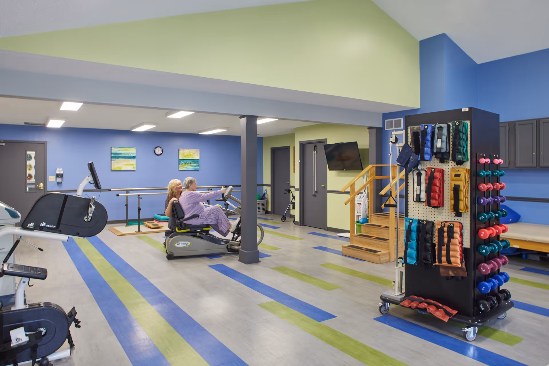 A senior woman exercises on a NuStep recumbent cross trainer with assistance from a caregiver in a brightly lit physical therapy or rehabilitation room. The room has blue and green striped flooring, blue and green walls, exercise equipment including weights and resistance bands, a wall-mounted TV, and a small set of stairs with a handrail.