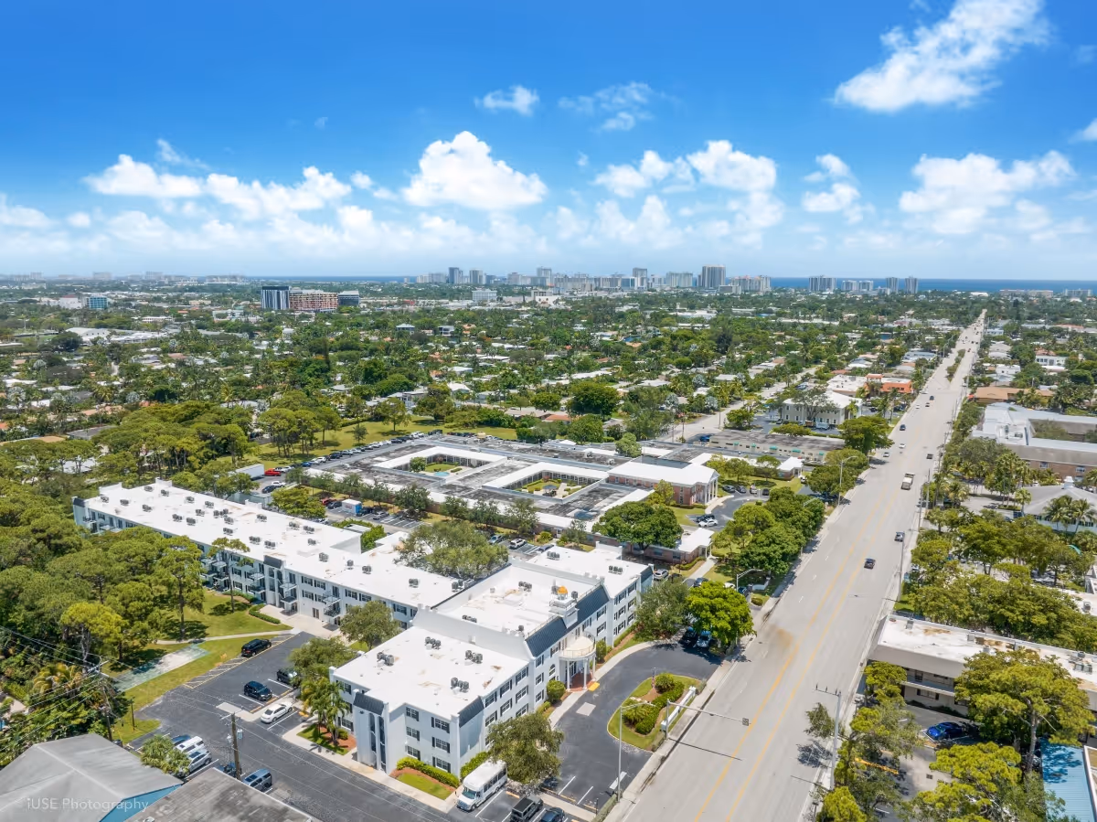 Aerial view of Independence Hall Senior Living facility surrounded by trees and roads, with a long street extending into the distance under a blue sky with scattered clouds.