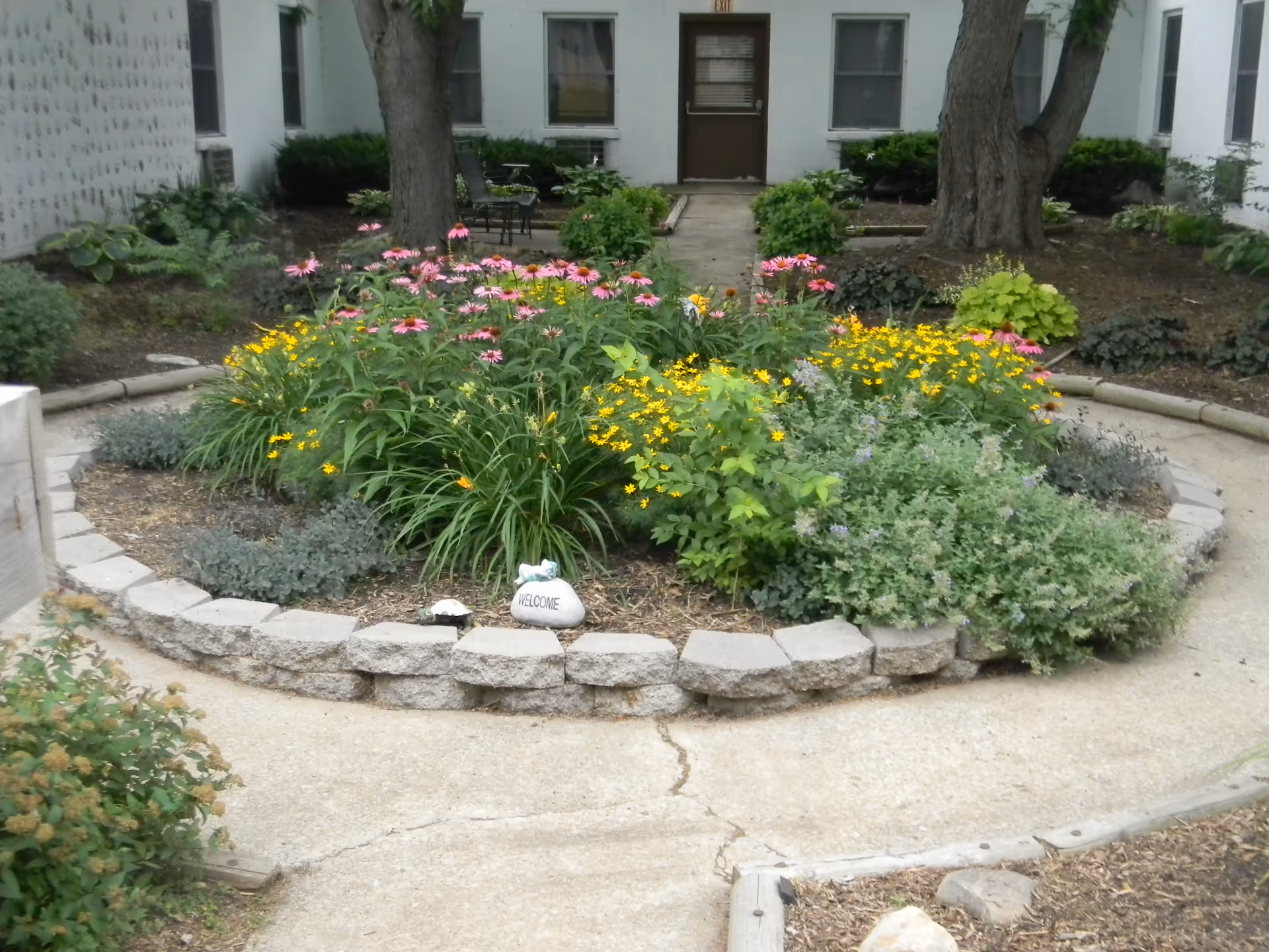 A circular garden bed with various flowering plants including pink and yellow flowers, surrounded by a low stone border. The garden is located in an outdoor courtyard area with concrete pathways and trees, with a building and windows visible in the background. A small stone with the word 'WELCOME' is placed in the garden bed.