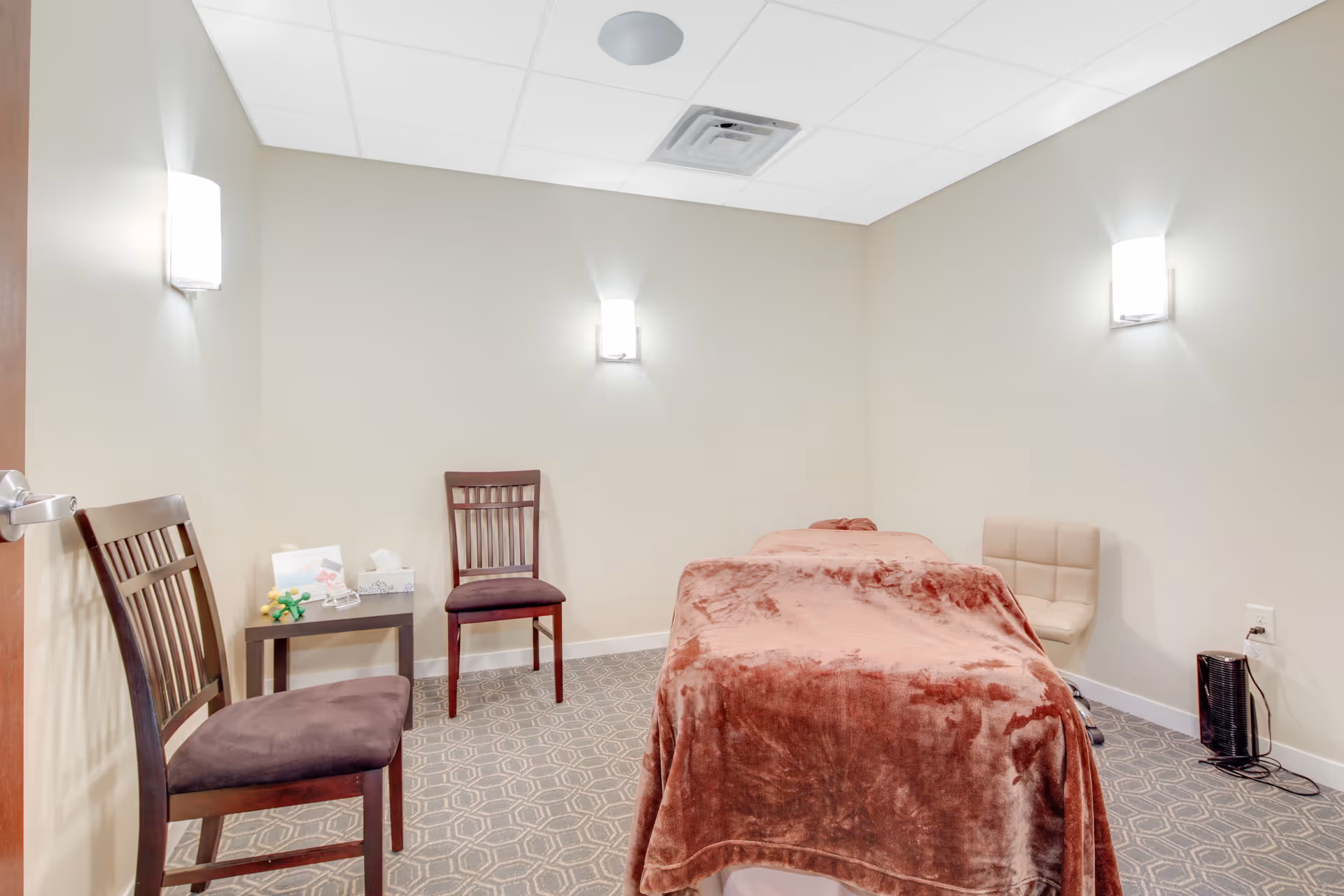 Small therapy/treatment room with a massage table covered by a brown blanket, seating chairs, and soft wall lighting.