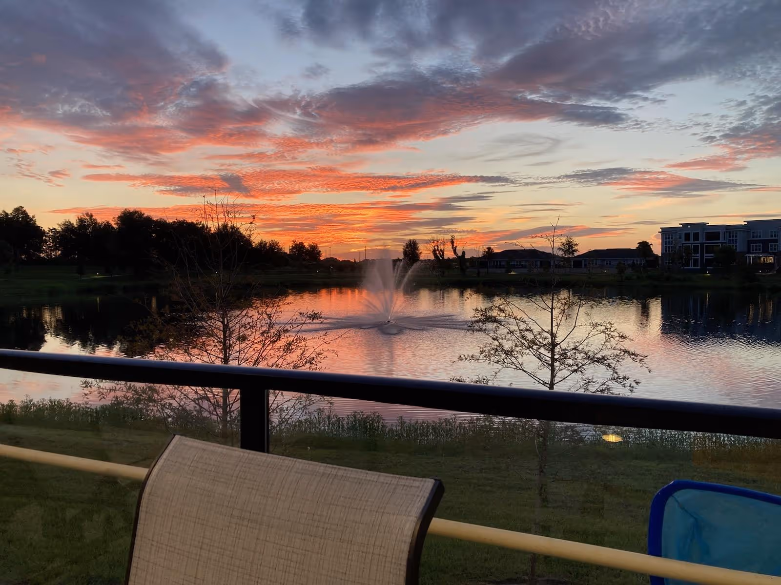 Sunset over a lake with a central fountain, seen from a balcony with a railing and a chair in the foreground.