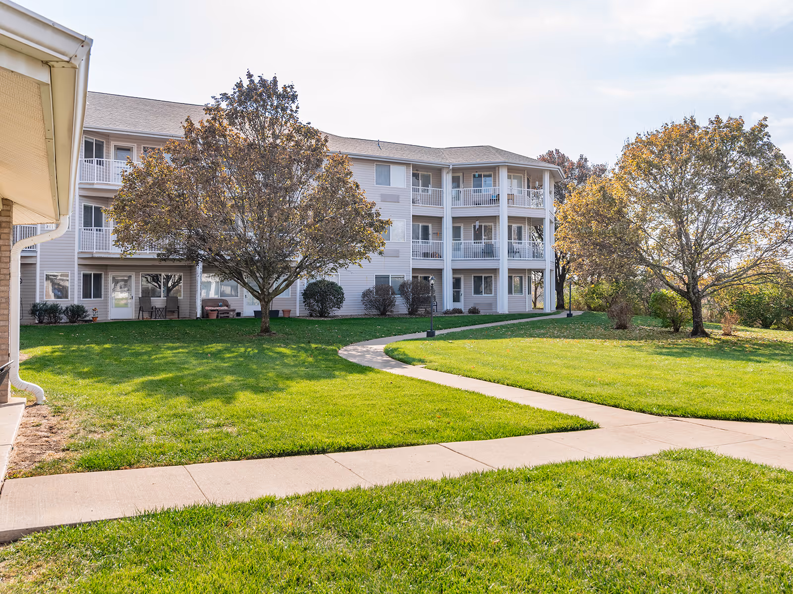 Exterior view of a three-story residential building with balconies, surrounded by green grass, trees with autumn foliage, and a curved concrete walkway leading to the entrance.