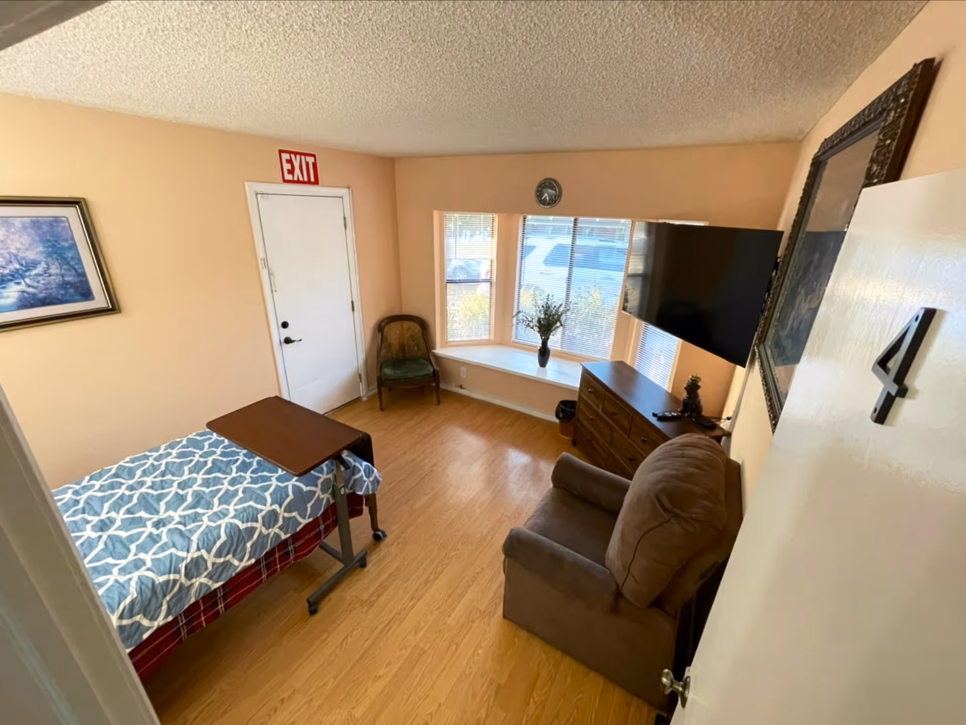A small, cozy room with a single bed covered with a blue and white patterned blanket, a brown recliner chair, a wooden dresser with a flat-screen TV mounted above it, a green cushioned chair in the corner, and a window with blinds and a vase of flowers on the windowsill. The walls are painted light peach, and there is an exit door with a red exit sign above it.