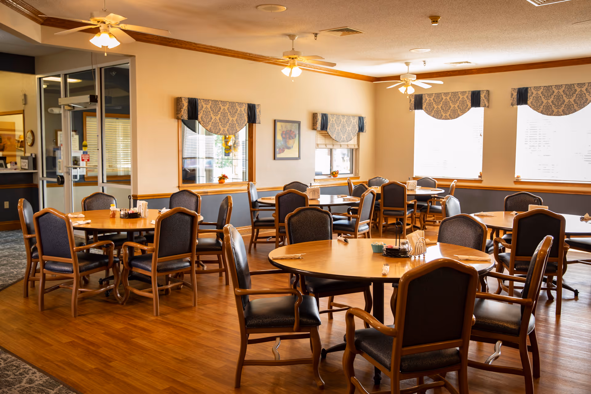 A well-lit dining room with multiple round wooden tables and cushioned chairs arranged neatly. The room features large windows with patterned valances, ceiling fans with lights, and a wooden floor. There are small decorative items and condiments on the tables.