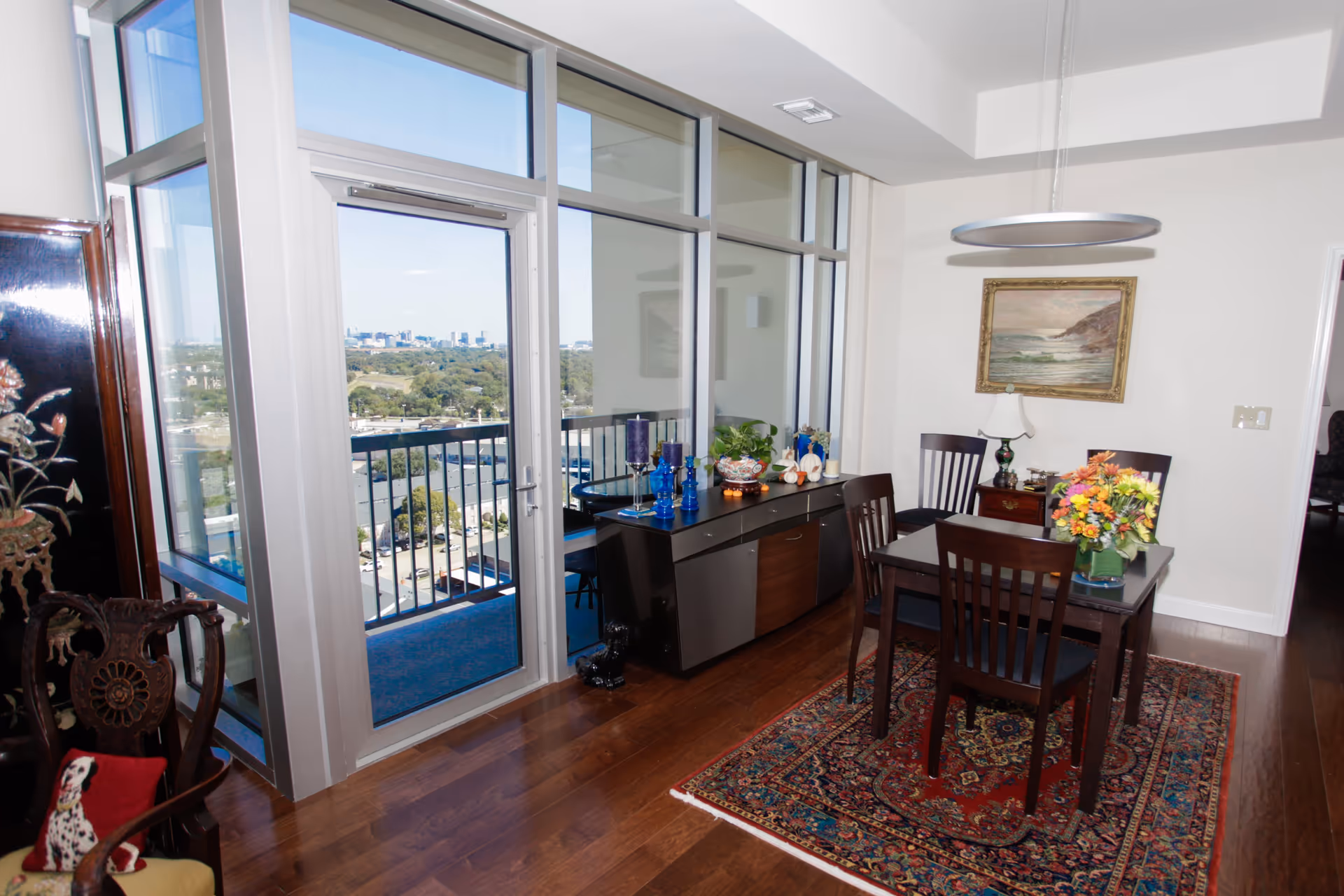 A dining area with a dark wooden table and four chairs on a patterned rug. A sideboard with decorative items including blue candles and a plant is against the wall. Large floor-to-ceiling windows and a glass door open to a balcony with a view of a city skyline and greenery. A framed painting and a modern circular light fixture are visible on the wall and ceiling.