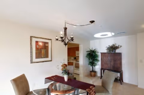 Bright dining area with a round glass table and chairs, a chandelier overhead, framed artwork on the wall, and a view into a kitchen with a decorative cabinet and potted plant.