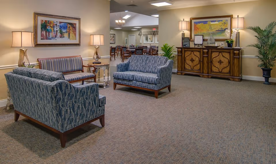 A cozy senior living facility common area with two blue patterned sofas and a striped loveseat arranged around a small side table with a lamp. The room is warmly lit with wall lamps and table lamps, decorated with framed paintings and a large wooden sideboard. In the background, there is a dining area with tables and chairs.