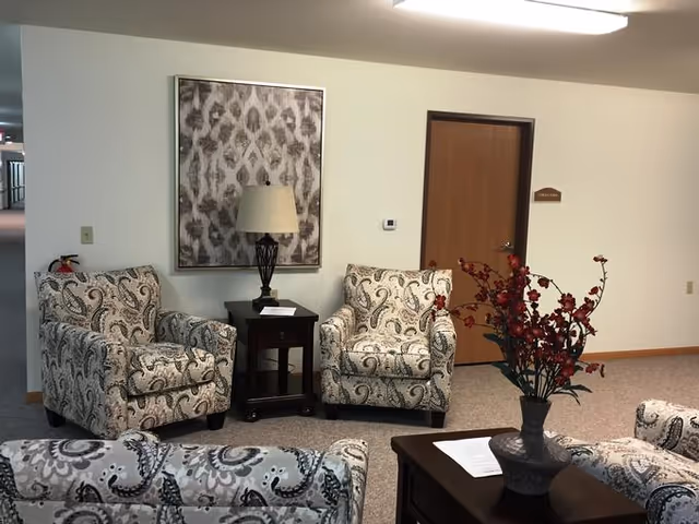 A cozy sitting area with patterned armchairs arranged around a wooden coffee table with a vase of red flowers. A side table with a lamp and a framed abstract artwork are against the wall. A wooden door and a hallway are visible in the background.