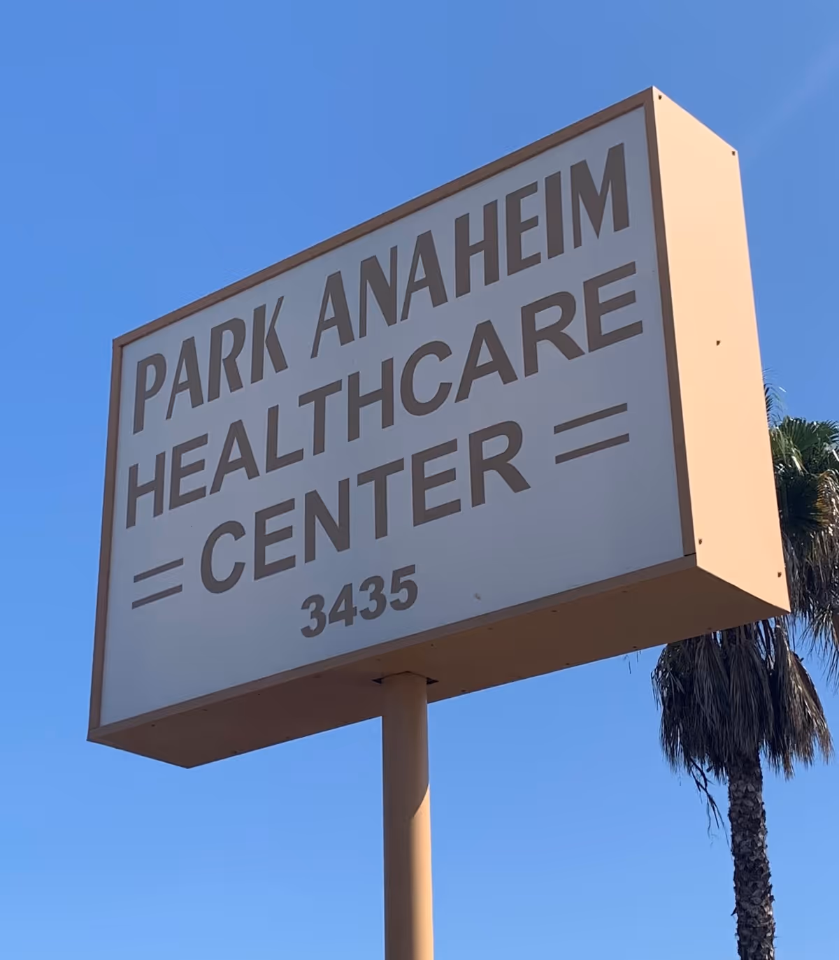 A large rectangular sign for Park Anaheim Healthcare Center mounted on a tall pole against a clear blue sky, with a palm tree visible in the background.