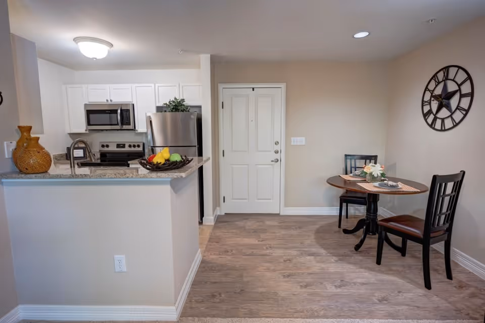 Interior view of a senior living facility apartment showing a small dining area with a round wooden table and two chairs, a decorative wall clock, and a kitchen with white cabinets, stainless steel appliances, and a granite countertop with a fruit basket and decorative vases.