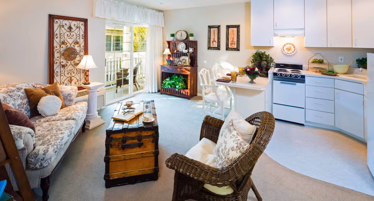 A cozy living area in an assisted living community featuring a floral patterned sofa with pillows, a wicker chair with cushions, a wooden trunk used as a coffee table, and a small dining counter with two white chairs. The space includes a compact kitchen with white cabinets, a stove, and a sink. A sliding glass door with white curtains leads to an outdoor balcony. Decorative items such as framed artwork, a lamp, plants, and a wooden shelf with plates and other decor complete the room.