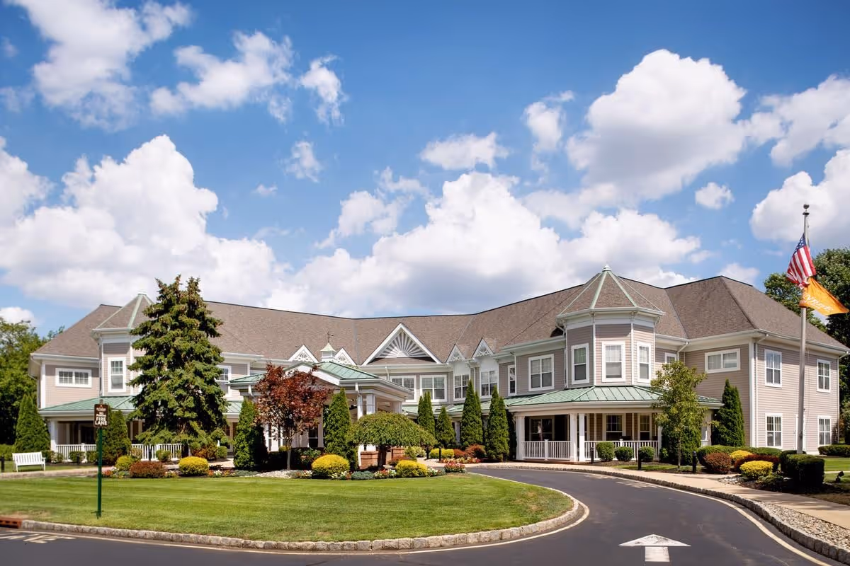 Exterior view of a large, two-story senior living facility building with beige siding and green metal roofing accents. The building is surrounded by well-maintained landscaping including bushes, trees, and a green lawn. A curved driveway leads to the entrance, and an American flag and an orange flag are flying on a flagpole to the right. The sky is blue with scattered white clouds.