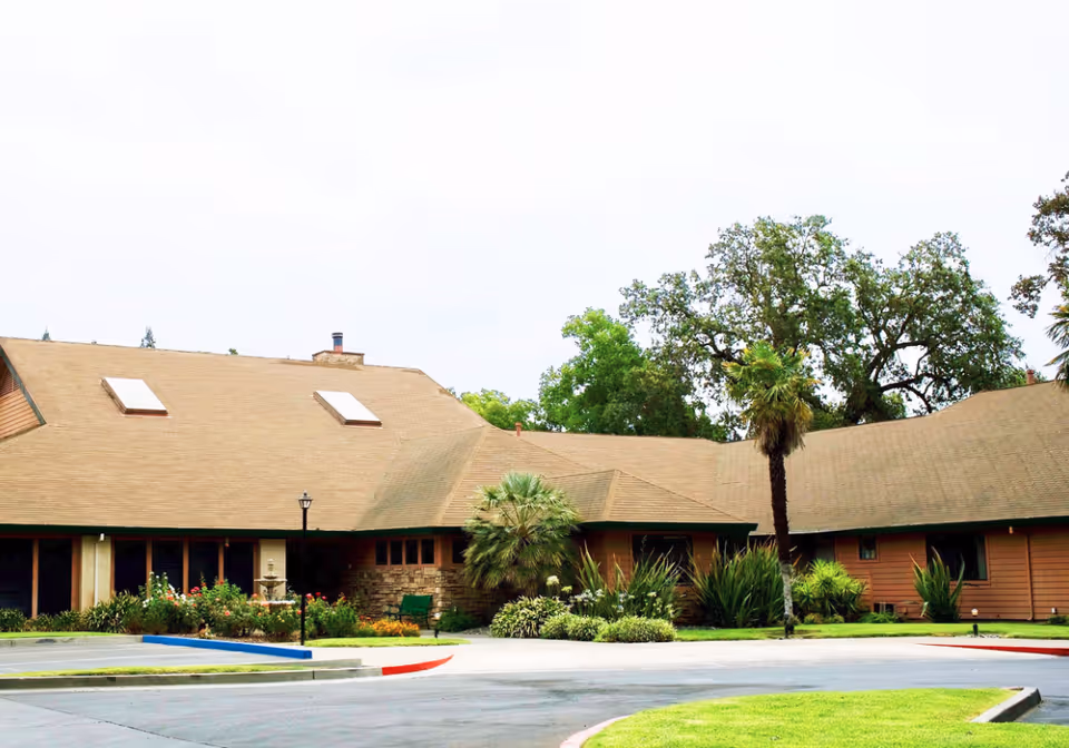 Front exterior of a single-story senior living building with a wide brown roof, landscaped gardens, palm trees, and a circular driveway.