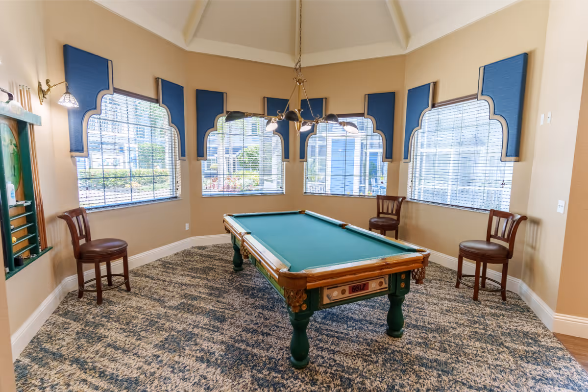 A bright interior game room featuring a central pool table under a hanging light with chairs around large arched windows.