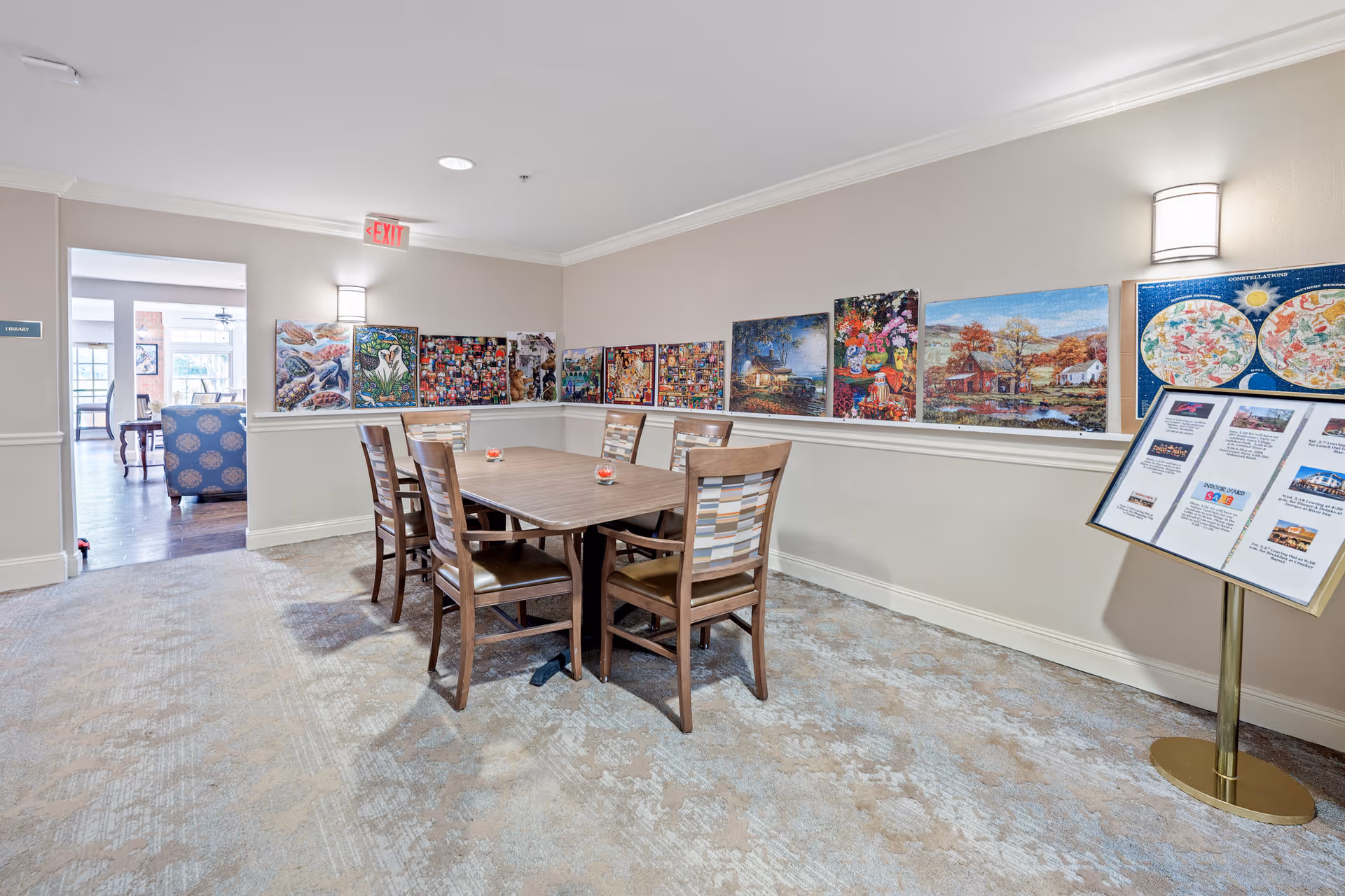 A well-lit room with a wooden table and six chairs arranged around it. The walls are decorated with colorful artwork and paintings. There is a stand with informational pamphlets or notices on the right side of the image. The room has beige walls, carpeted floor, and a doorway leading to another room with additional seating visible.