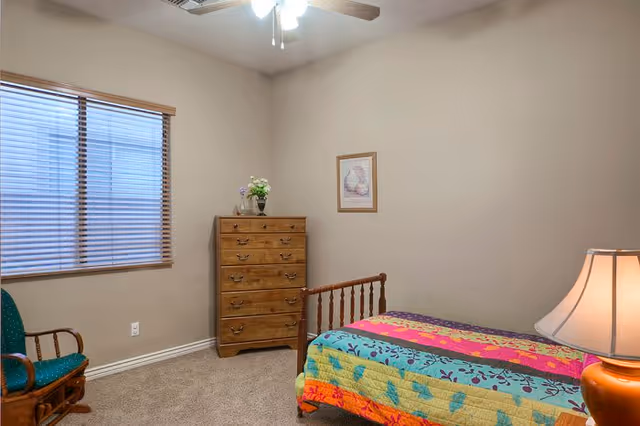 A small bedroom with beige walls and carpeted floor featuring a wooden bed with a colorful quilt, a wooden chest of drawers with a vase of flowers on top, a framed picture on the wall, a green cushioned rocking chair, and a window with wooden blinds. A ceiling fan with light is visible above.