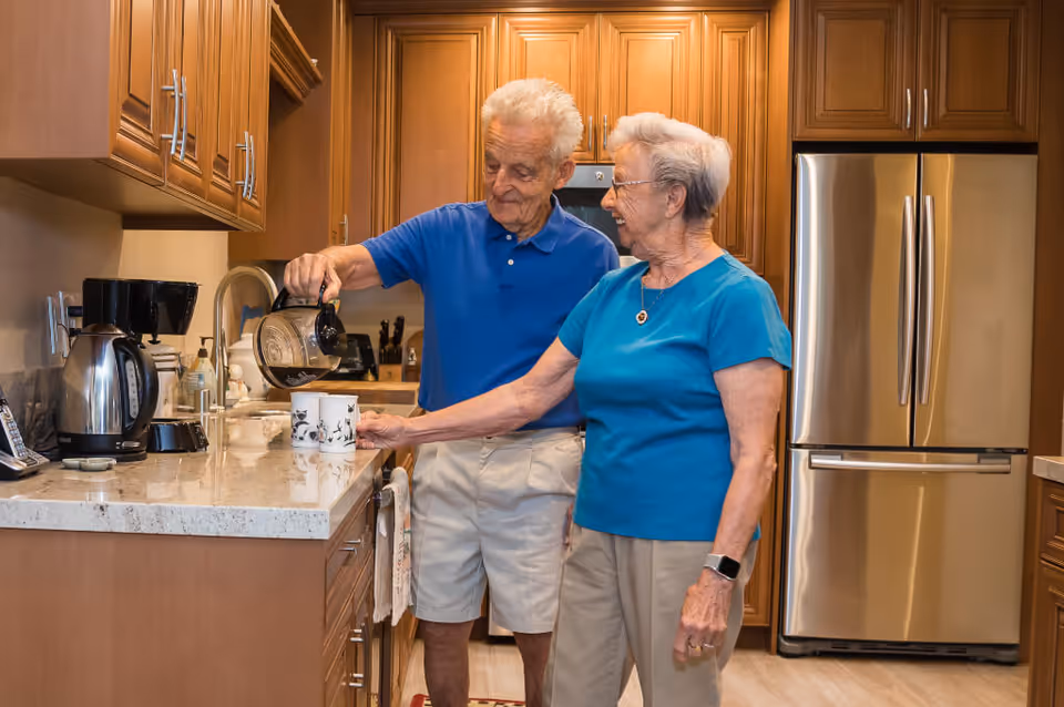 An elderly man is pouring coffee into a mug held by an elderly woman in a kitchen with wooden cabinets and a stainless steel refrigerator. Both are smiling and appear to be enjoying the moment.