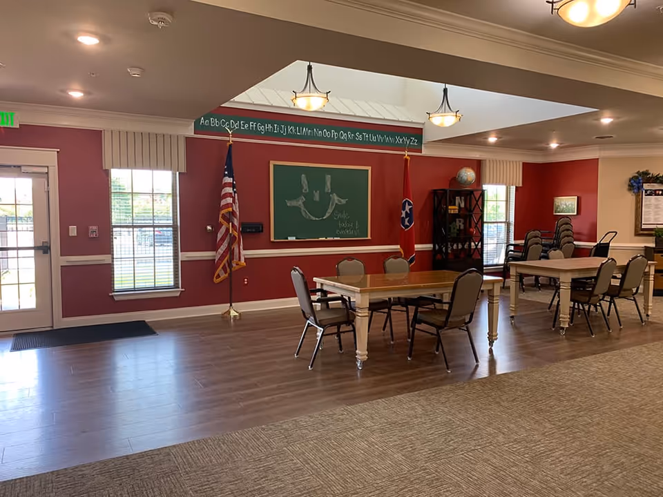 Interior of a community room with red walls and large windows. There are two tables with chairs around them, an American flag and a Tennessee state flag, a chalkboard with a smiley face and the words 'Smile, laugh & enjoy life!', and a bookshelf with a globe and decorative letters. The room has wood flooring and carpet, with ceiling lights and a door leading outside.