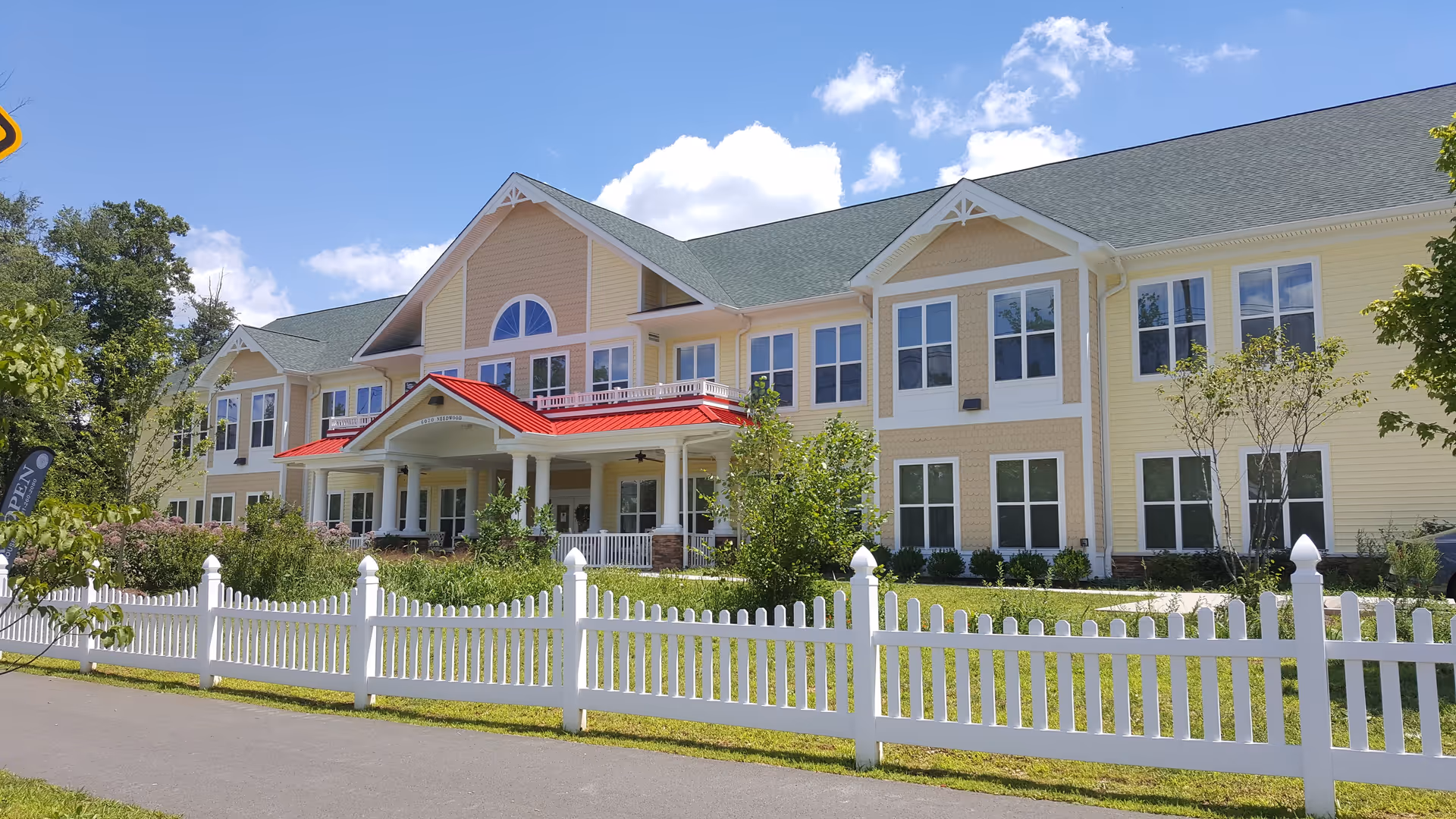 Exterior view of AlfredHouse Symphony, a two-story senior living facility with a light yellow facade, green roof, and a red awning over the entrance. The building is surrounded by greenery and a white picket fence under a blue sky with some clouds.