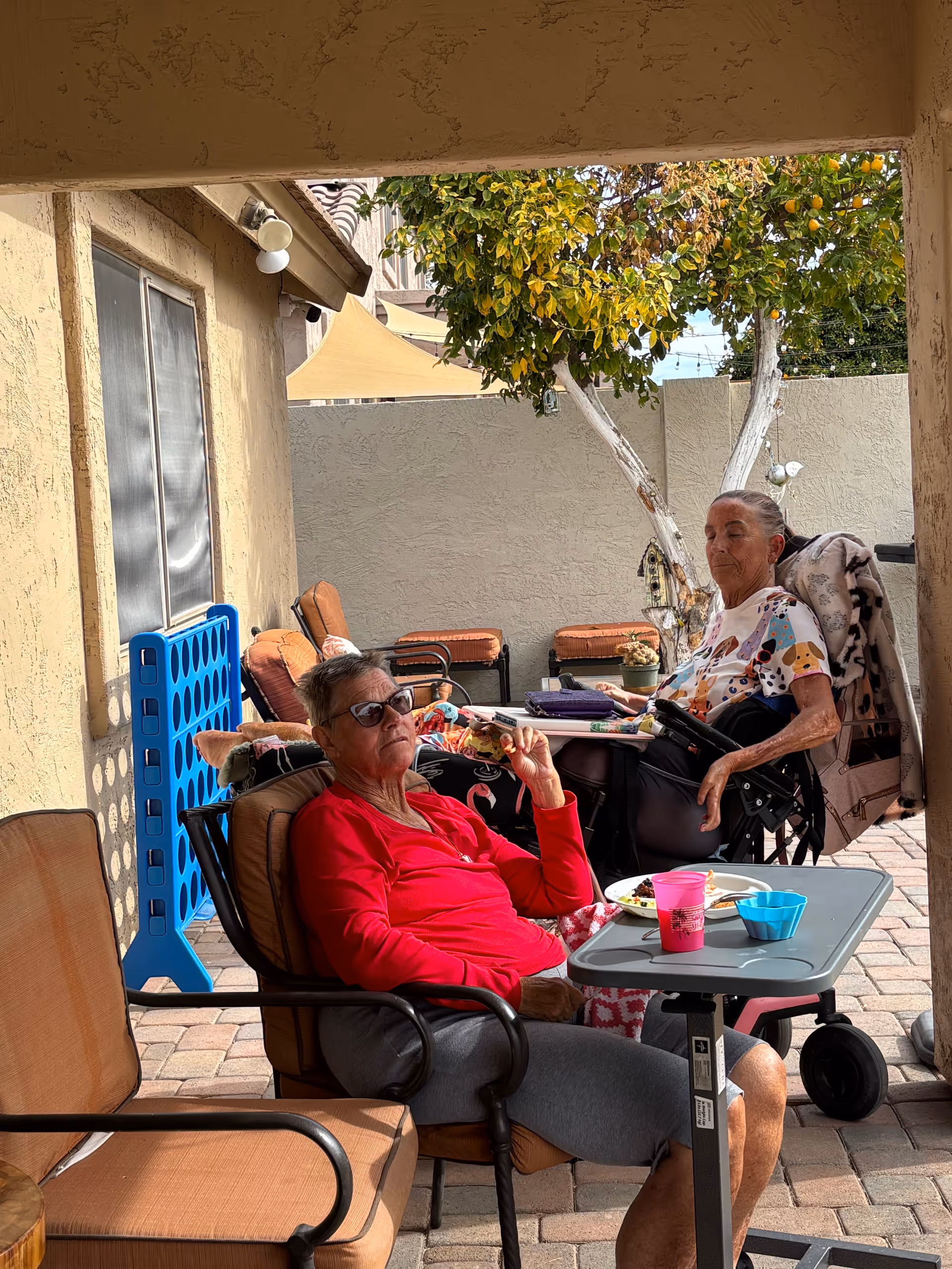Two elderly women seated on a shaded outdoor patio, one in a wheelchair, with patio chairs, a small table with cups, and a nearby tree.