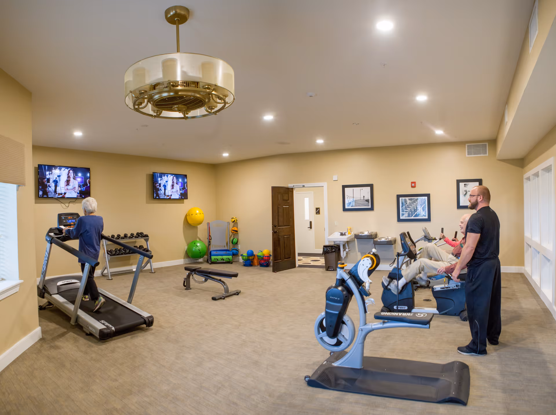 A bright indoor fitness room with treadmills, stationary bikes, exercise equipment, and residents using the machines.