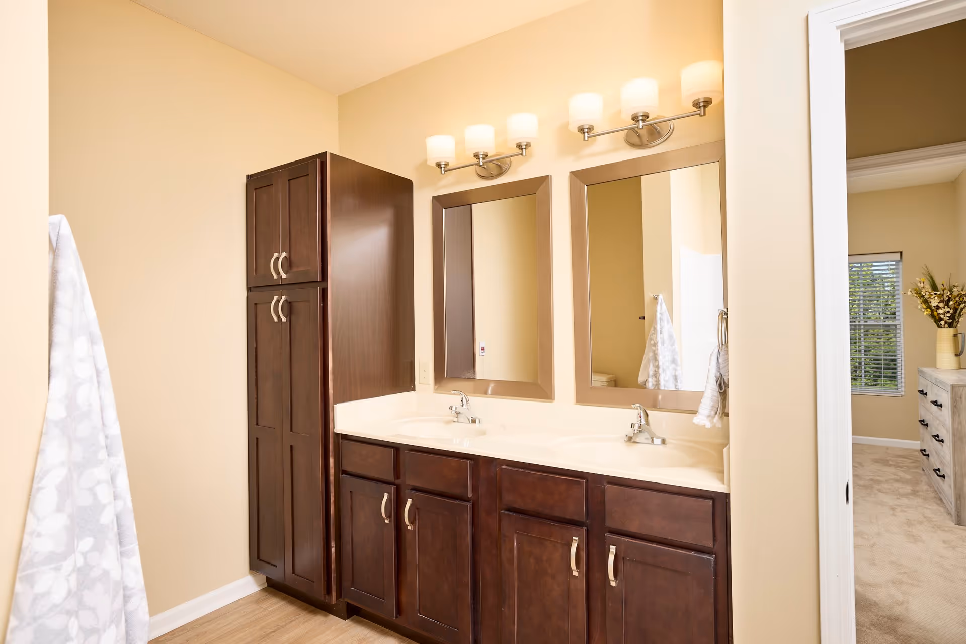 A bathroom with a double sink vanity featuring dark wood cabinets and two mirrors above. There are two light fixtures with four lights each mounted above the mirrors. To the left of the vanity is a tall dark wood cabinet. A towel hangs on a hook on the left wall. Through an open doorway on the right, a bedroom with a window and a dresser with a vase of flowers is visible.
