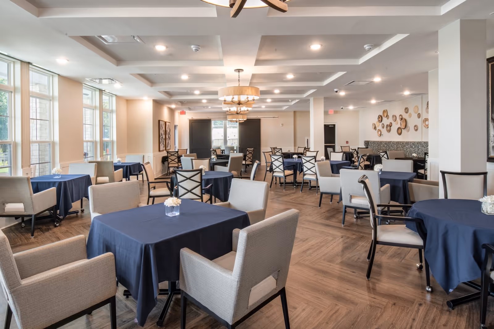 A spacious dining room with multiple tables covered in dark blue tablecloths, surrounded by beige and black chairs. The room has large windows on one side allowing natural light to enter, modern ceiling lights, and decorative wall art. The floor is wooden with a herringbone pattern.