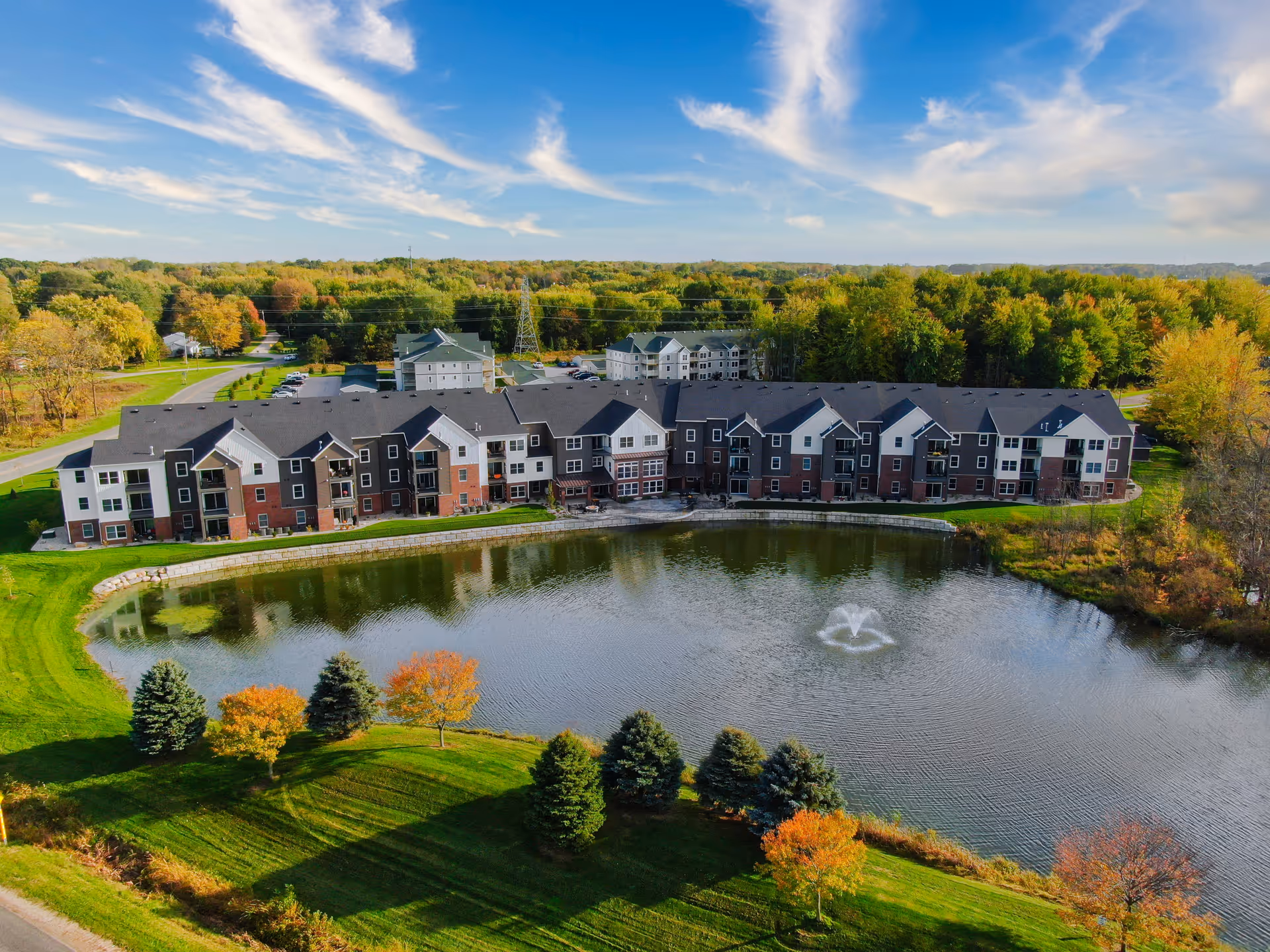 Aerial view of a senior living facility named Kensington Senior Living, featuring a large multi-story building with a mix of dark and light exterior colors, situated beside a pond with a water fountain. The surrounding area includes green lawns, trees with autumn foliage, and a clear blue sky with scattered clouds.
