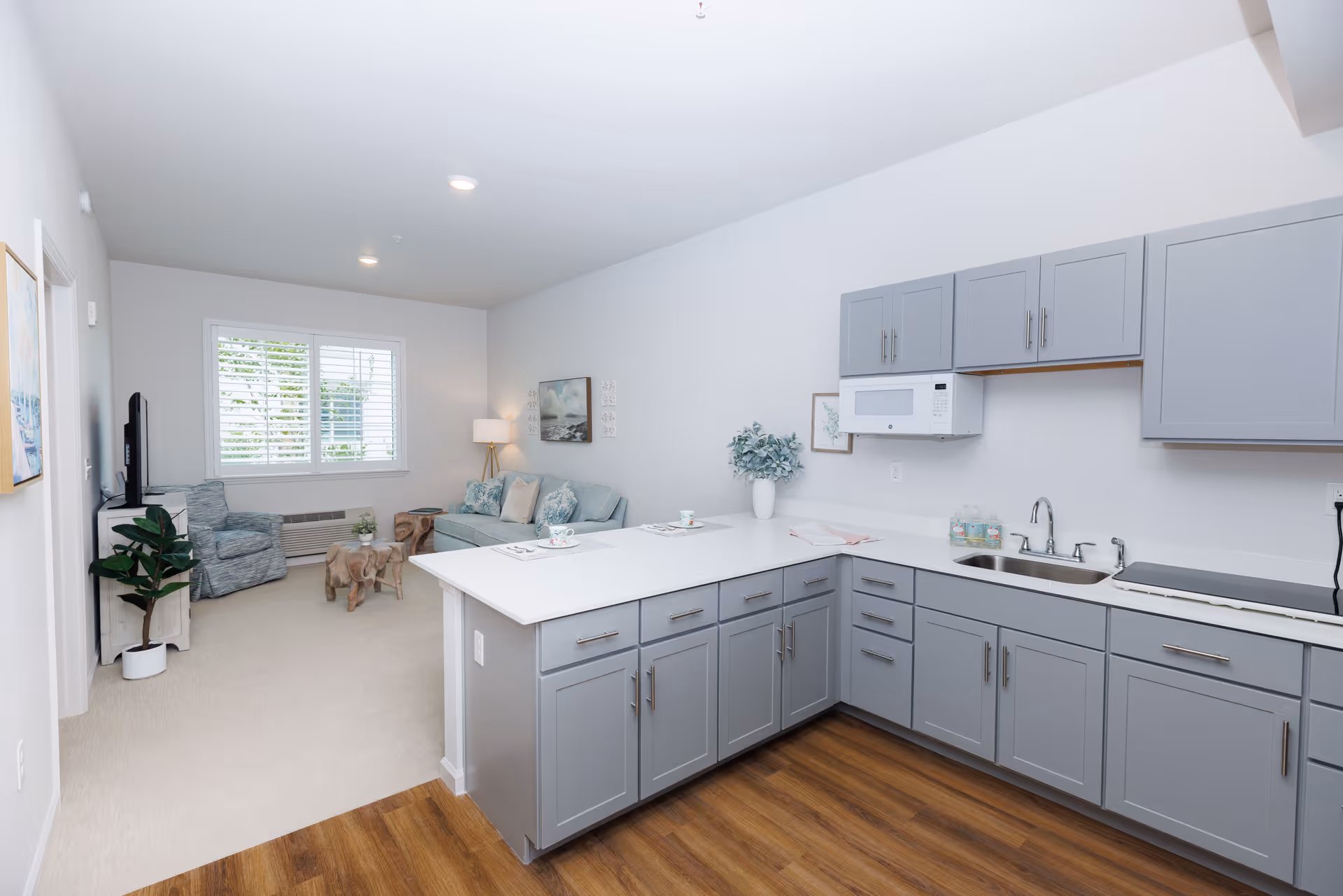 A bright and modern senior living apartment interior featuring a kitchen with gray cabinets, white countertops, a microwave, and a sink. The kitchen opens into a living area with a light blue sofa, an armchair, a wooden coffee table, a floor lamp, and a window with white shutters letting in natural light.