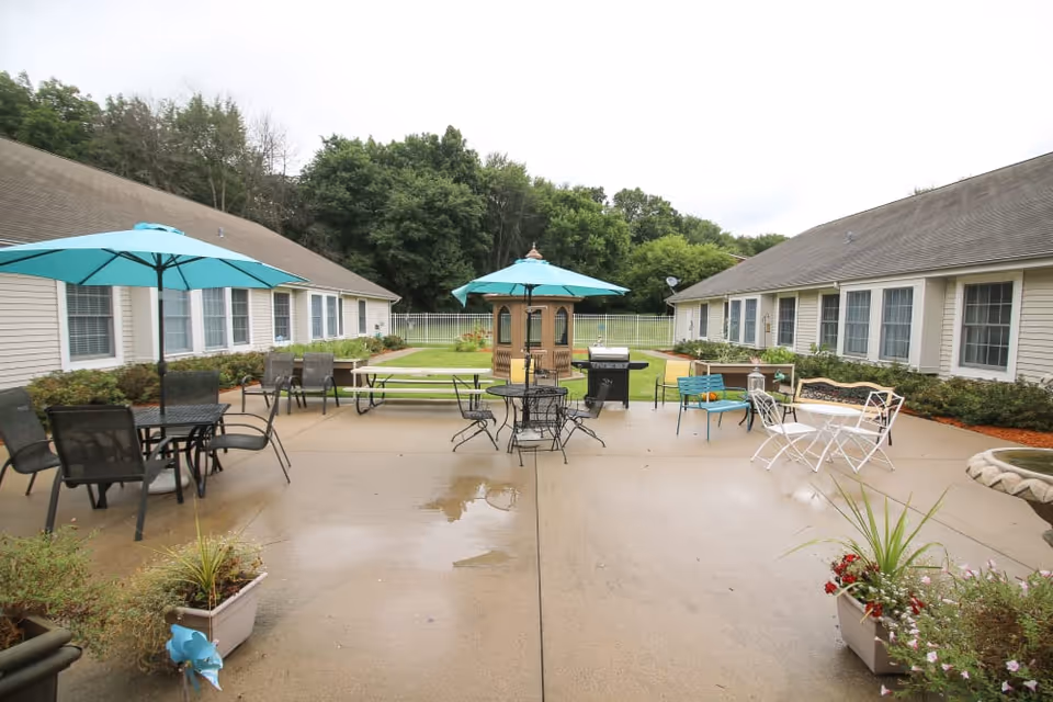 Outdoor patio area between two single-story buildings with beige siding and multiple windows. The patio has several tables and chairs, some with turquoise umbrellas, a picnic table, a small gazebo, a grill, and various potted plants. Trees and a white fence are visible in the background.