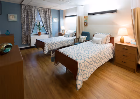 Two neatly made twin beds in a well-lit senior living bedroom with nightstands, a lamp, a chair, a dresser, and a window with patterned curtains.