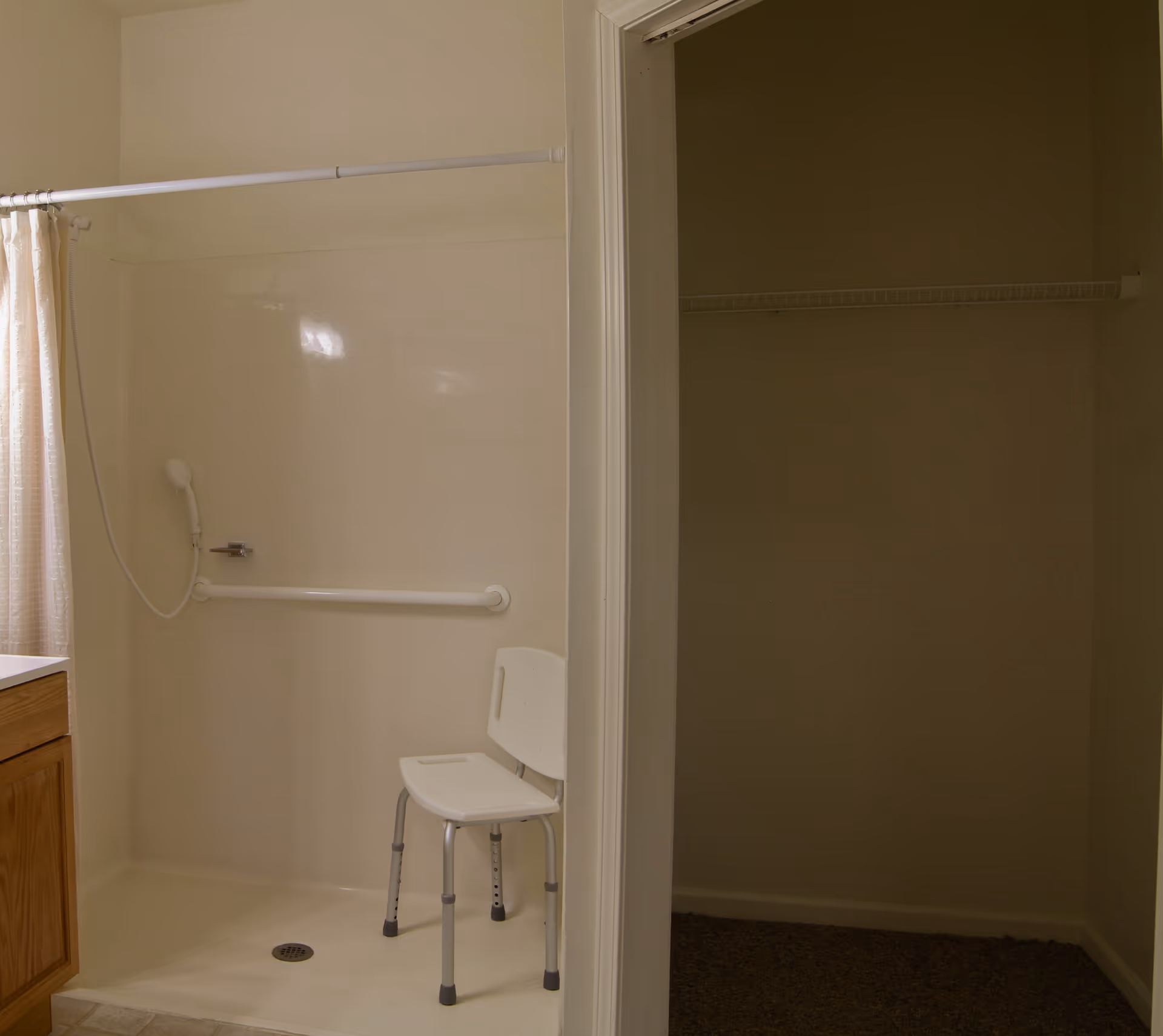 A bathroom shower area with a white shower chair, a handheld showerhead, and a grab bar mounted on the wall. To the right of the shower is an empty closet with a wire shelf. The bathroom has light-colored walls and a wooden cabinet partially visible on the left.