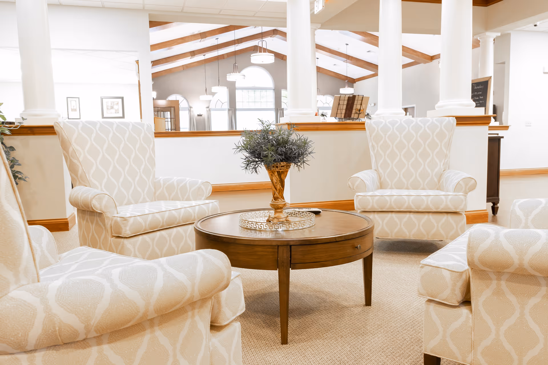 A bright and airy senior living common area with four patterned beige armchairs arranged around a round wooden coffee table. The table has a decorative gold vase with greenery and a small tray. The background shows white columns, large windows, and hanging light fixtures with wooden beams on the ceiling.