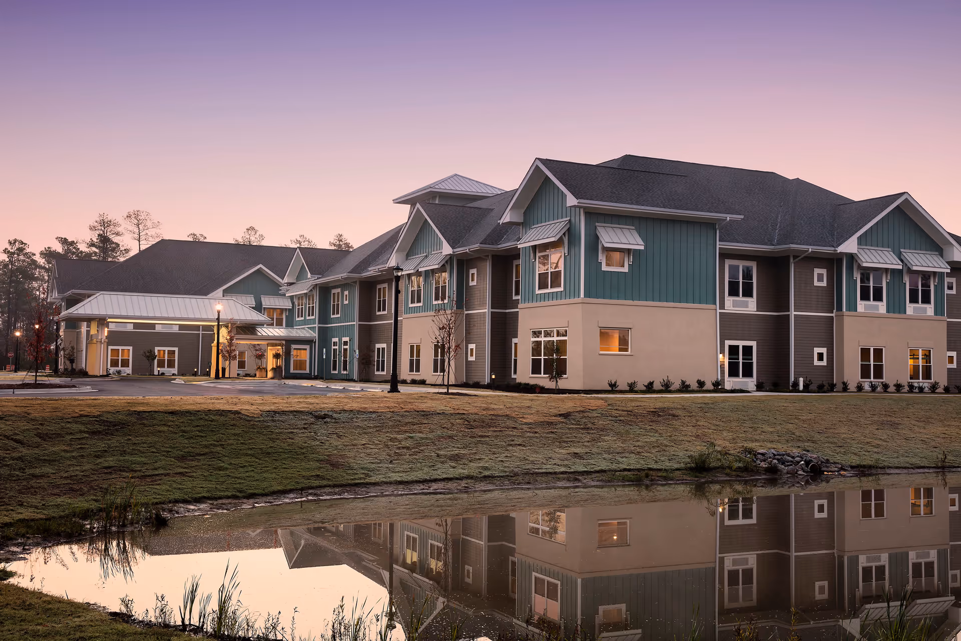Exterior view of a two-story senior living facility building at dusk with a calm pond in the foreground reflecting the building. The building has a combination of teal and beige siding with multiple windows and a covered entrance. The sky is a soft gradient of purple and pink.