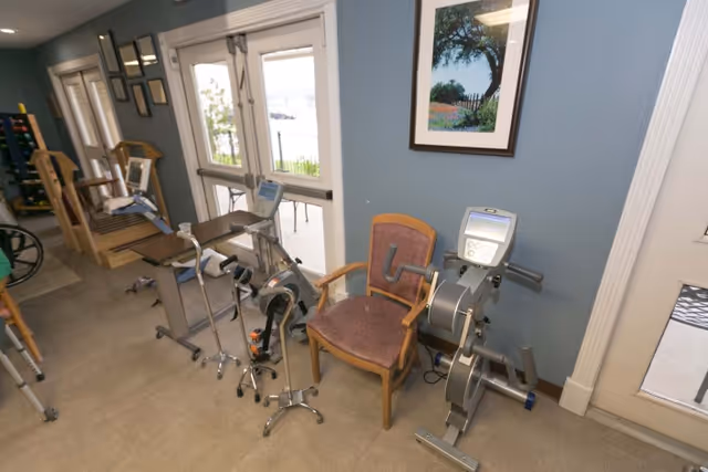 A rehabilitation room with exercise equipment including a stationary pedal exerciser, a chair, and other physical therapy tools. The room has blue walls, a framed picture of a tree, and glass doors leading outside.