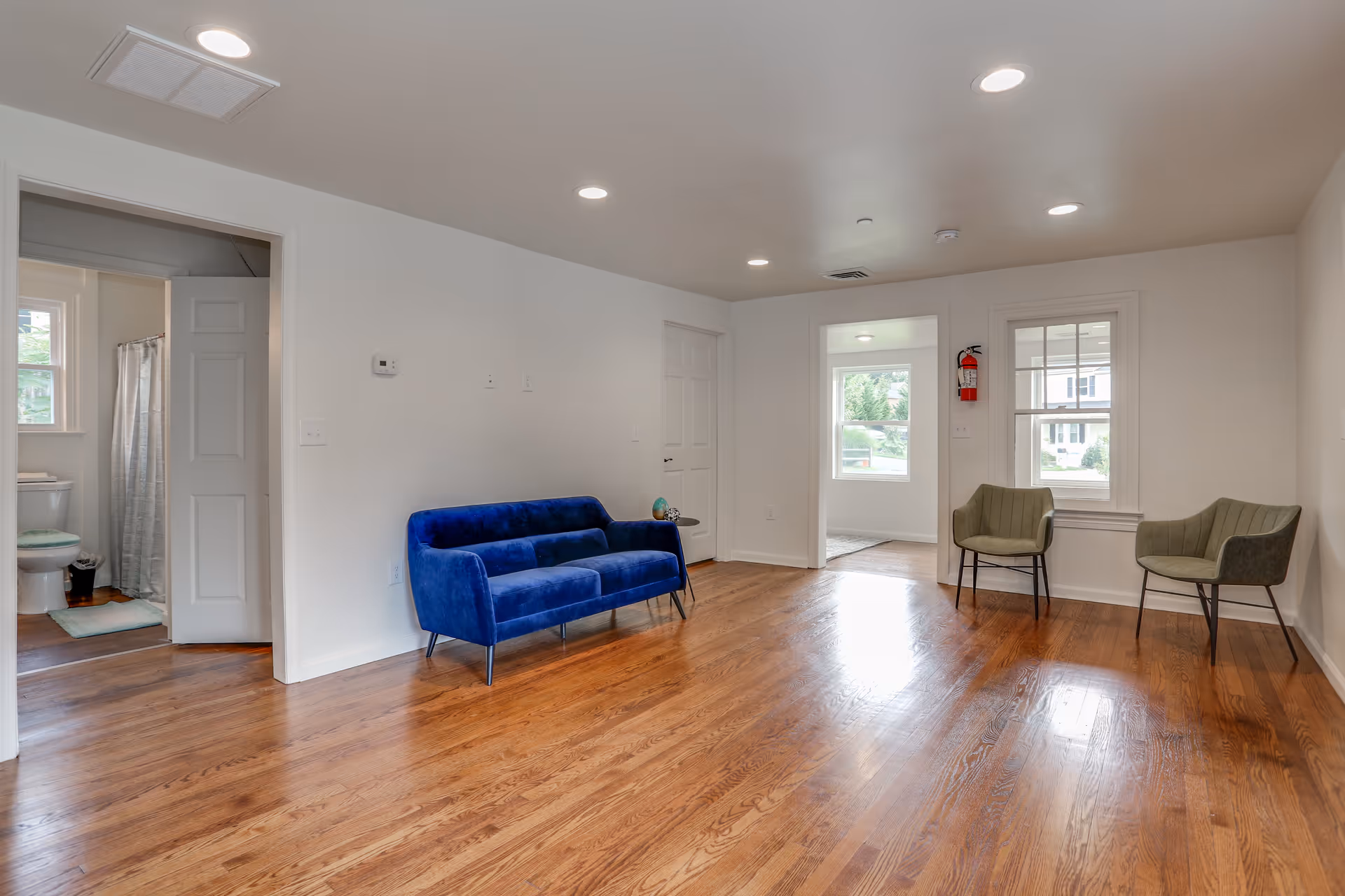 Open living room with hardwood floors, a blue velvet sofa and two green chairs by windows, with a bathroom visible through a doorway on the left.