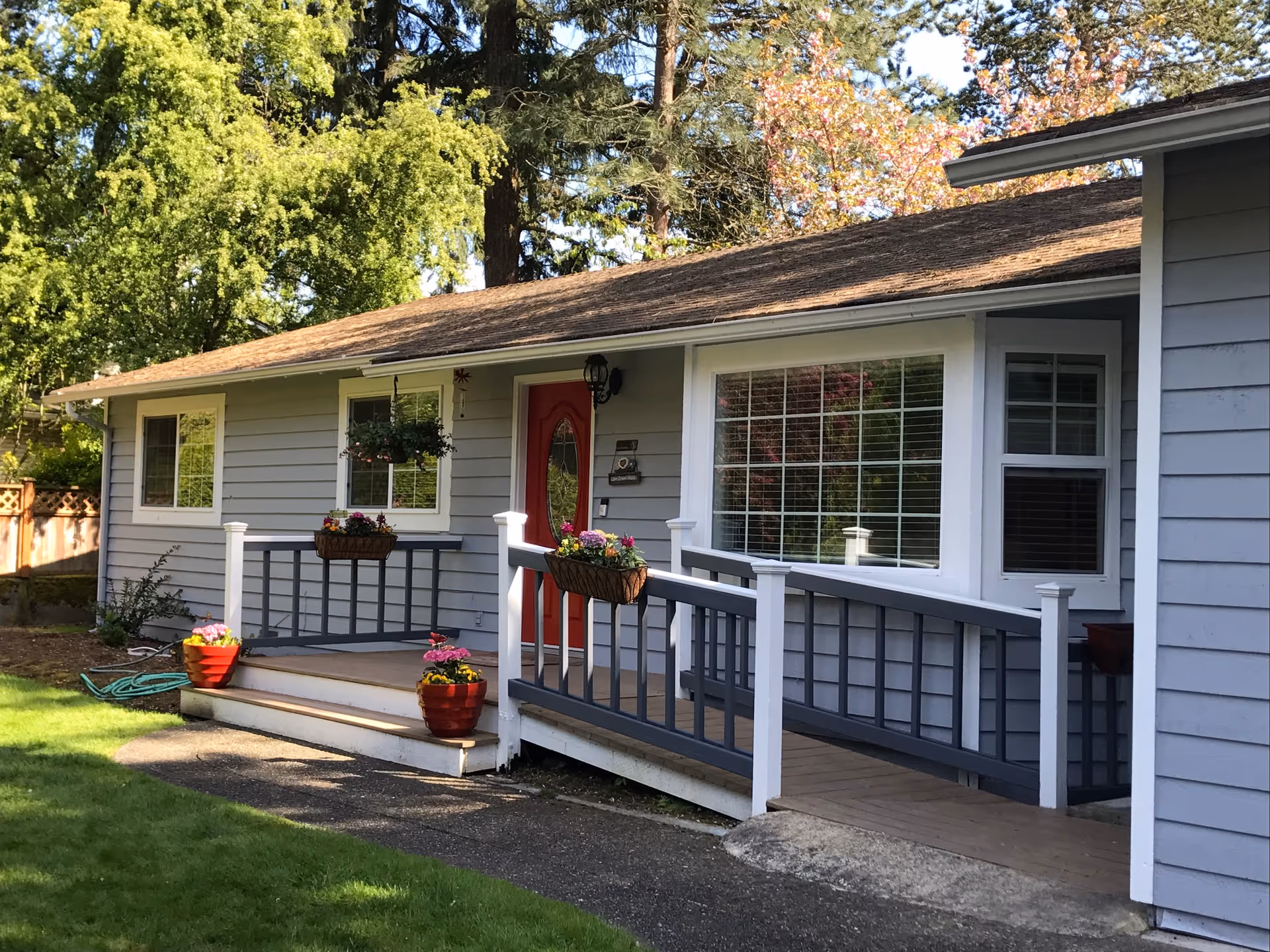 Exterior view of a single-story gray house with white trim and a red front door. The house has a wooden ramp and steps leading to the entrance, decorated with flower pots and hanging baskets. The surrounding area includes green grass, trees, and a wooden fence.