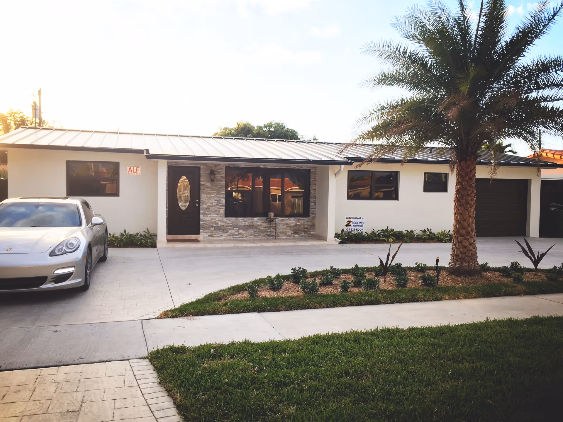 Front exterior view of a single-story building with a metal roof, a dark brown door with an oval window, and several windows. There is a silver car parked on the left side driveway, a palm tree, and landscaped garden beds in front of the building. A sign on the building reads 'ALF'.