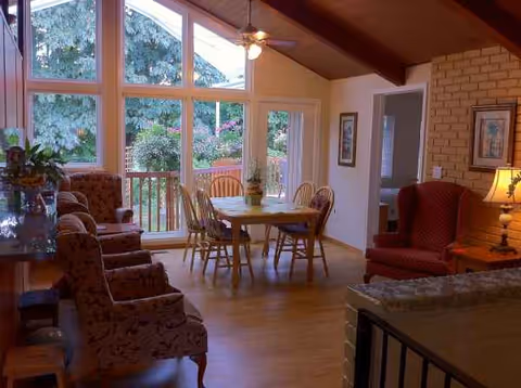 Sunlit senior living common area with large windows, wooden beams, brick fireplace, and comfy seating.